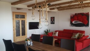 Et spiseområde med et træbord og stole, en rød sofa med puder med dyremotiver, et fjernsyn monteret på væggen, og et loft med synlige træbjælker og hængende lamper.