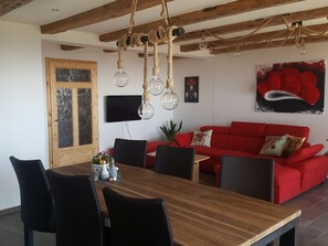 Uma sala de jantar com mesa e cadeiras de madeira, um sofá vermelho com almofadas de tema animal, uma TV na parede e um teto com vigas de madeira aparentes e luminárias pendentes.