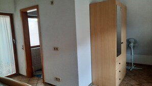 Una habitación con un armario de madera, una puerta que da a otra estancia y un ventilador de pie.