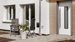 Un bâtiment moderne doté de grandes fenêtres, d’une porte avec un panneau en verre dépoli et d’un espace extérieur aménagé avec des chaises et un pot de fleurs.