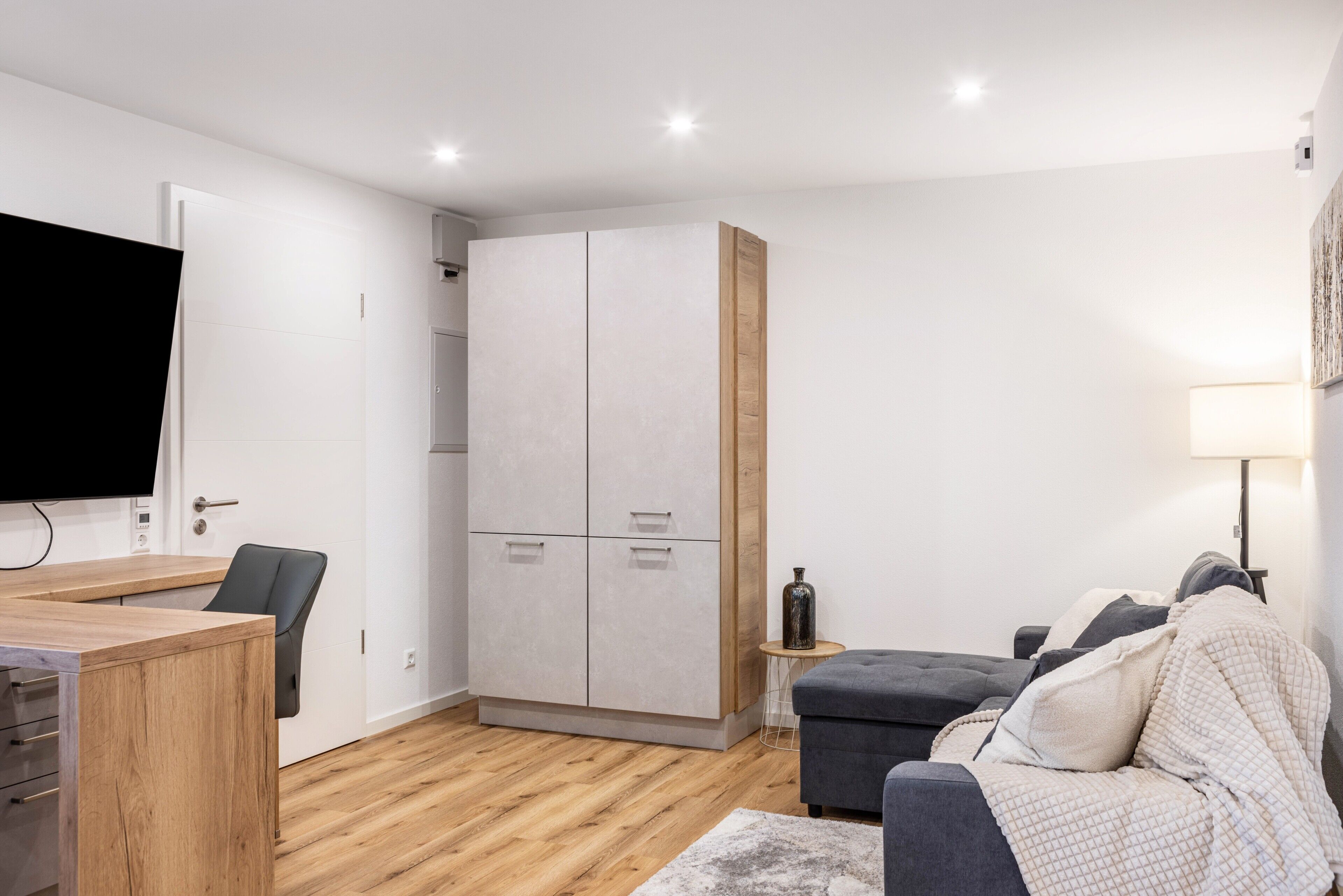 A modern room with a wooden desk, a grey sofa, a TV, and a wardrobe.