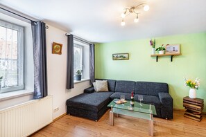Un salon comprenant un canapé, une table basse en verre, un radiateur, une fenêtre avec des rideaux, une horloge murale, un tableau encadré, une étagère avec des plantes et une petite table avec un vase.