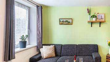 A living room with a sofa, a window with curtains, a small table with a bottle and glass, and a wall-mounted shelf with plants and a picture.