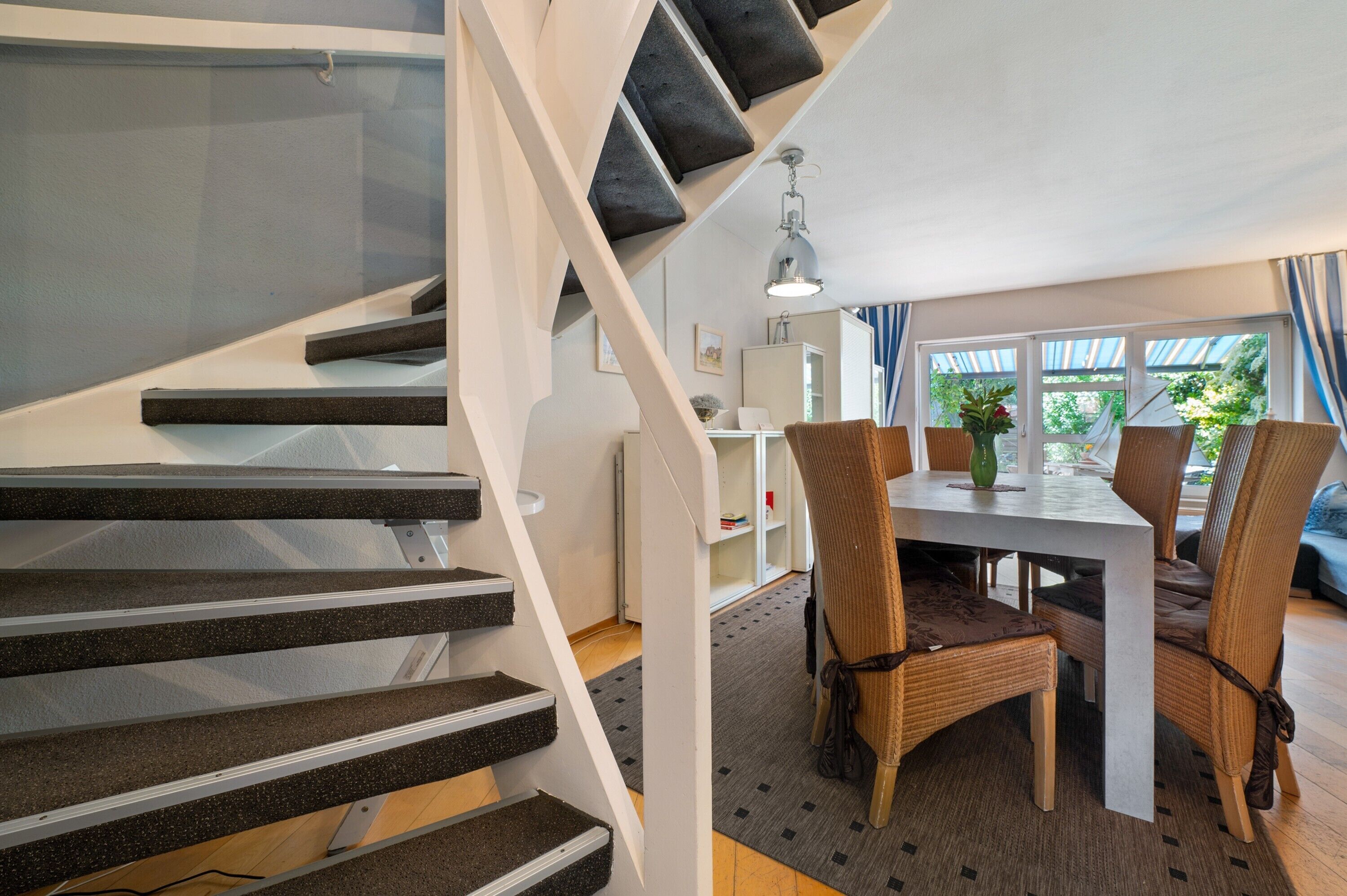 Un escalier au design moderne, un coin repas avec une table et des chaises, et une vue sur la verdure à l’extérieur.