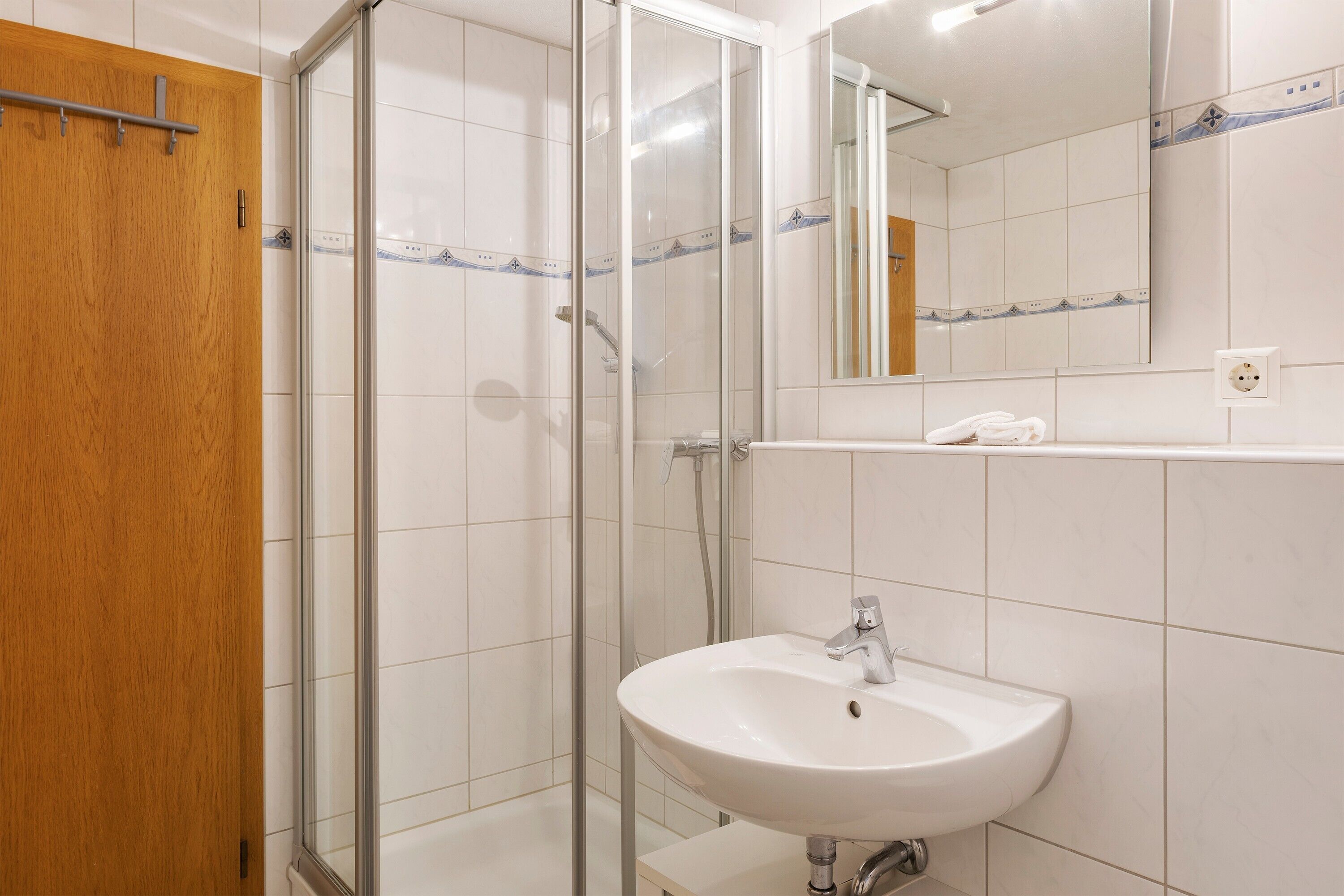A bathroom with a glass shower enclosure, a wooden door, a white sink with a mirror above it, and a tiled wall.