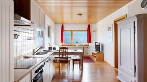A modern kitchen with wooden cabinets, a dining area with a table and chairs, a window with curtains, and a view of the outdoors.