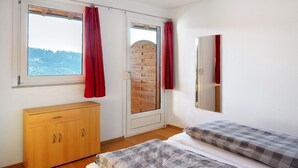 A bedroom with a wooden dresser, a bed with a checkered blanket, a window with curtains, and a brick wall.