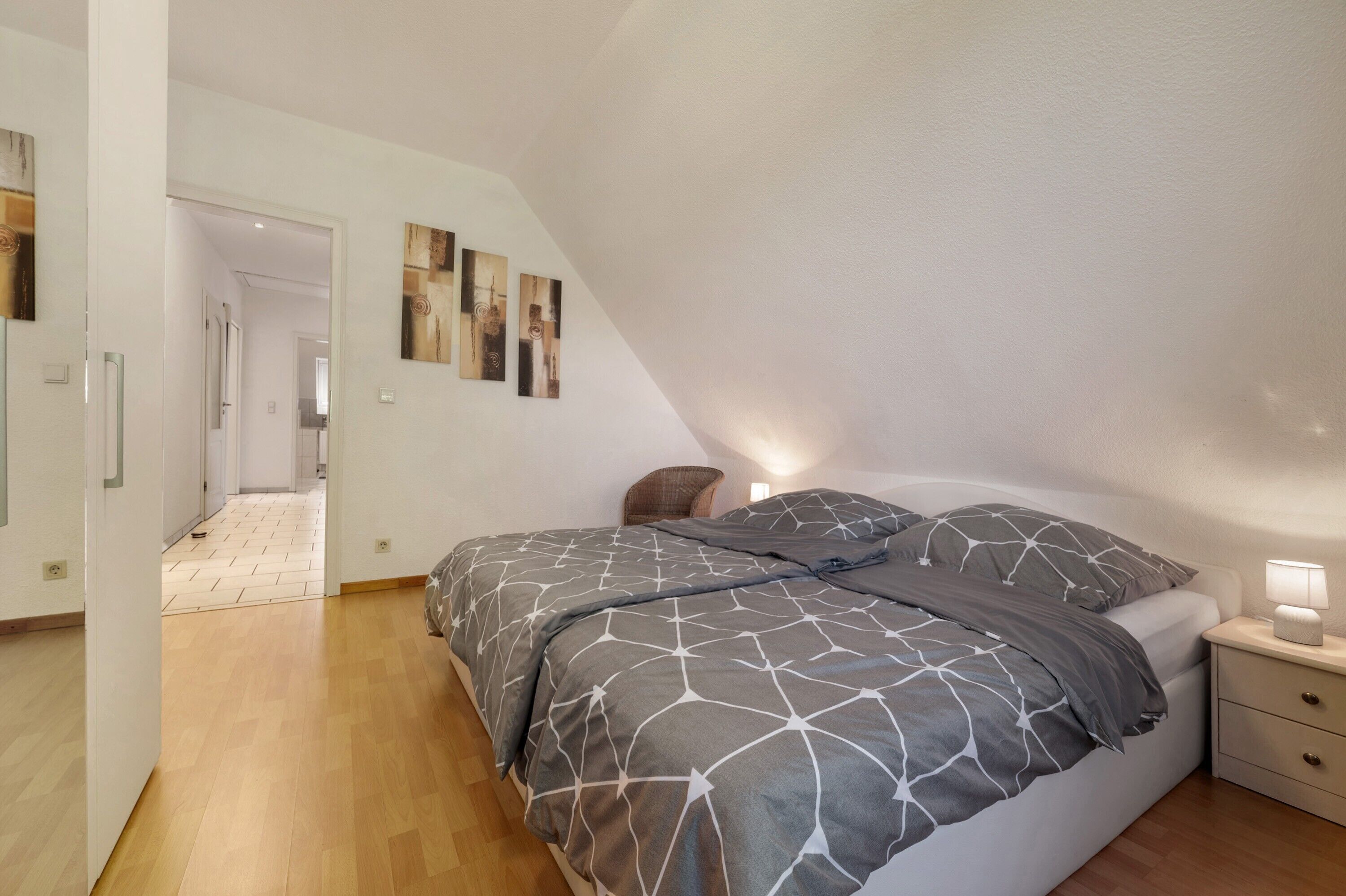 Una camera da letto con soffitto spiovente, un letto con copriletto a motivi geometrici, un comodino con una lampada e un corridoio che conduce a un'altra stanza.