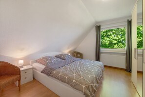 Ein Schlafzimmer mit Bett, Nachttisch, Stuhl, einem Fenster mit Vorhängen und Blick auf Grünfläche.