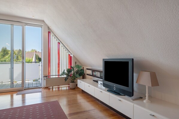 Un salon moderne avec un téléviseur, une lampe, une plante en pot et une porte-fenêtre coulissante donnant sur un balcon.