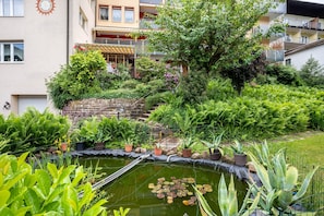Un jardín con un estanque, diversas plantas y un edificio con balcones.