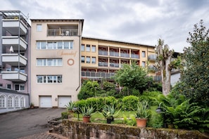 A multi-story hotel with balconies, a garden area with potted plants, and a sign with a sun symbol.