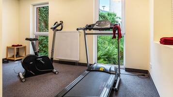 A fitness room with a treadmill, elliptical, and exercise bike, a radiator, and a window with a view of greenery.