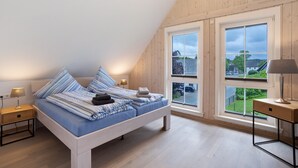 Un dormitorio con techo inclinado, una cama con ropa de cama azul, dos mesitas de noche con lámparas y ventanas amplias que ofrecen vistas al exterior.