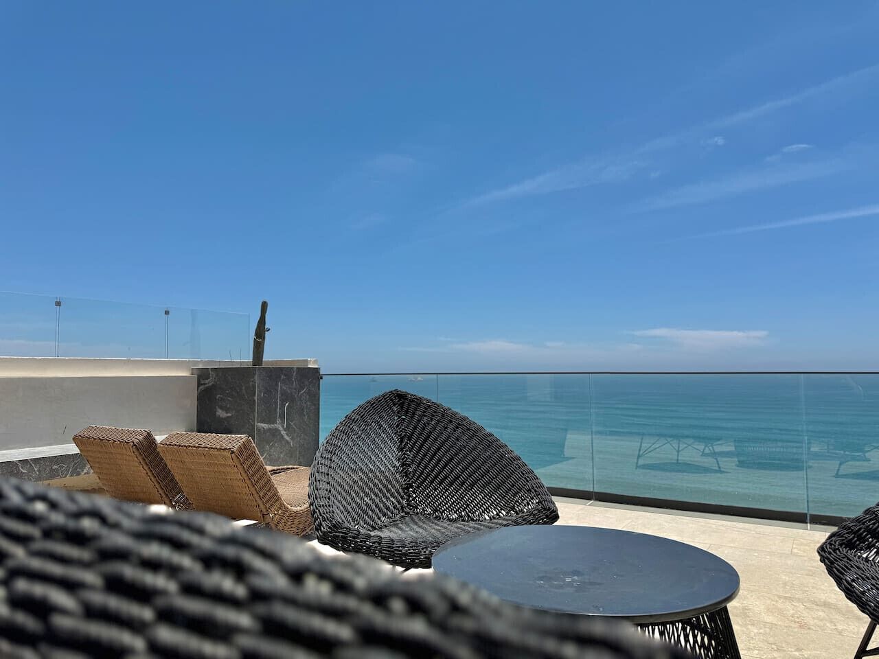 Un balcon avec des fauteuils en osier et une table, donnant sur une mer d’un bleu limpide.