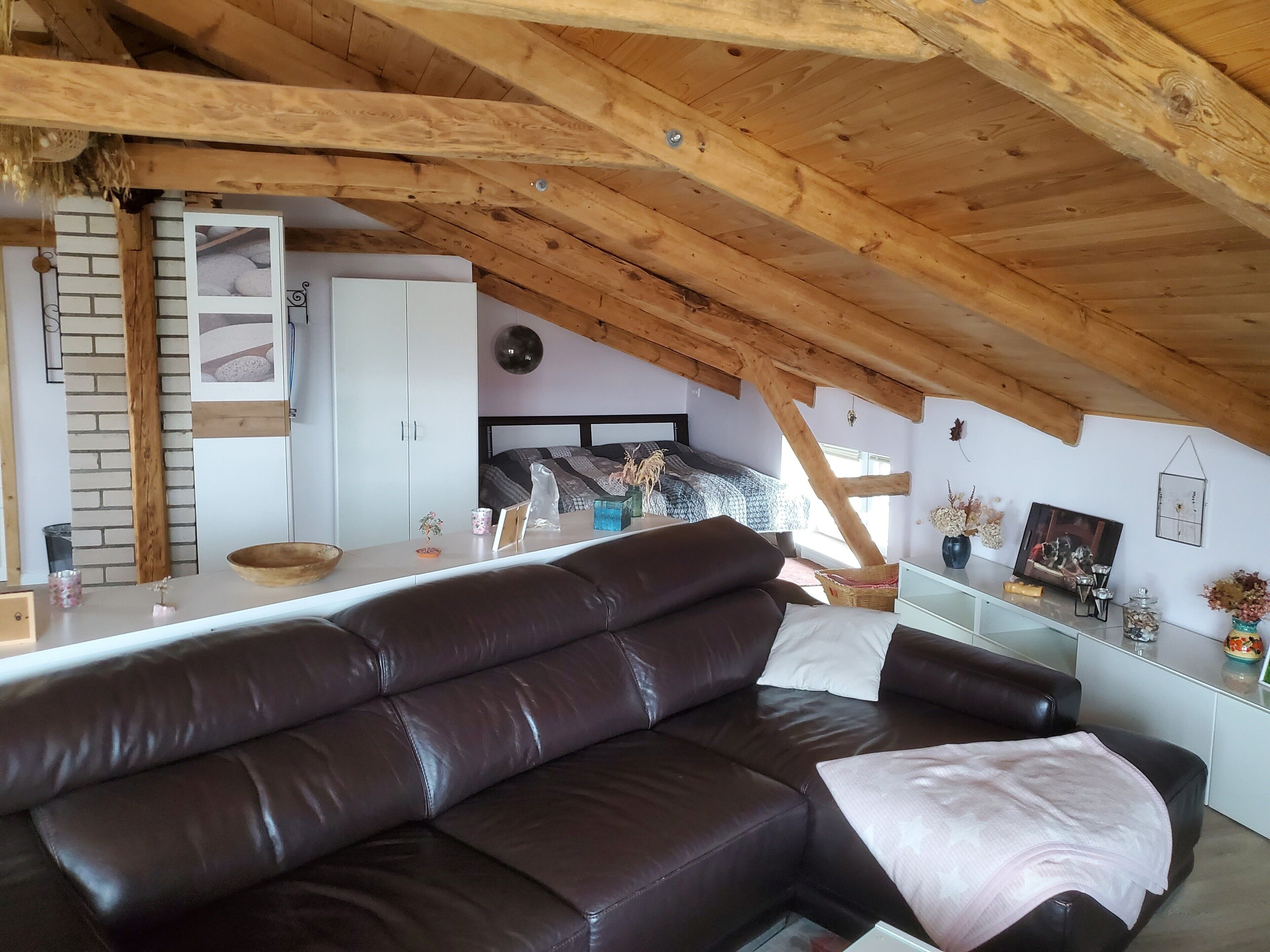 Avoin, loft-tyylinen huone, jossa on ruskea nahkasohva, kuviollinen päiväpeite, valkoinen vaatekaappi ja puinen katto.