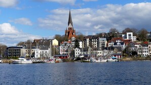 Een stadsgezicht aan het water met een opvallende kerktoren, woongebouwen en boten afgemeerd aan de oever.