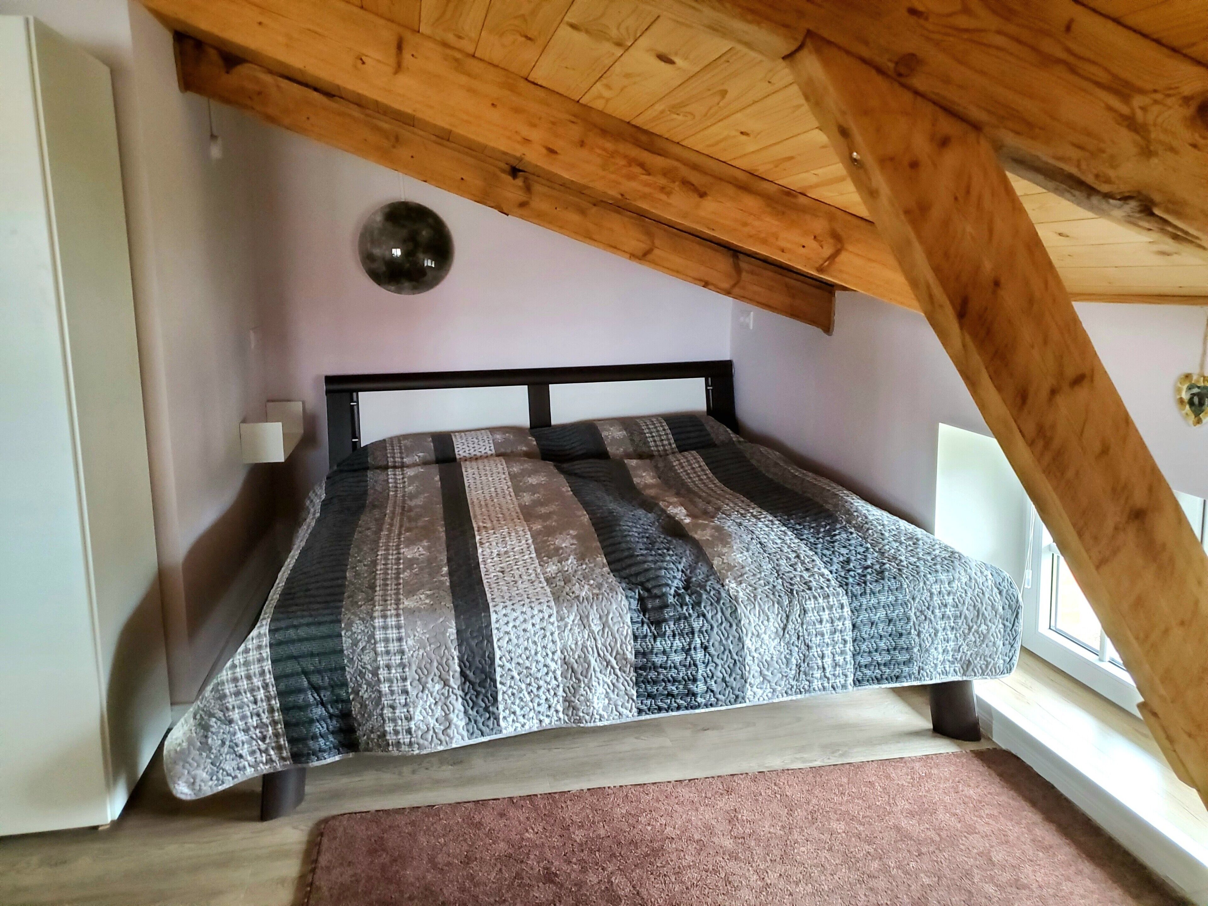 Ein Schlafzimmer mit schräg angesetzter Decke, einem Bett mit gemustertem Überwurf, einer Nachttischlampe und einem Fenster.