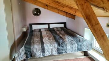 Ein Schlafzimmer mit schräg angesetzter Decke, einem Bett mit gemustertem Überwurf, einer Nachttischlampe und einem Fenster.