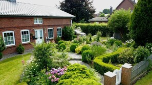 Een besloten tuin met een stenen huis, een verhard pad en verschillende struiken en bloemen.