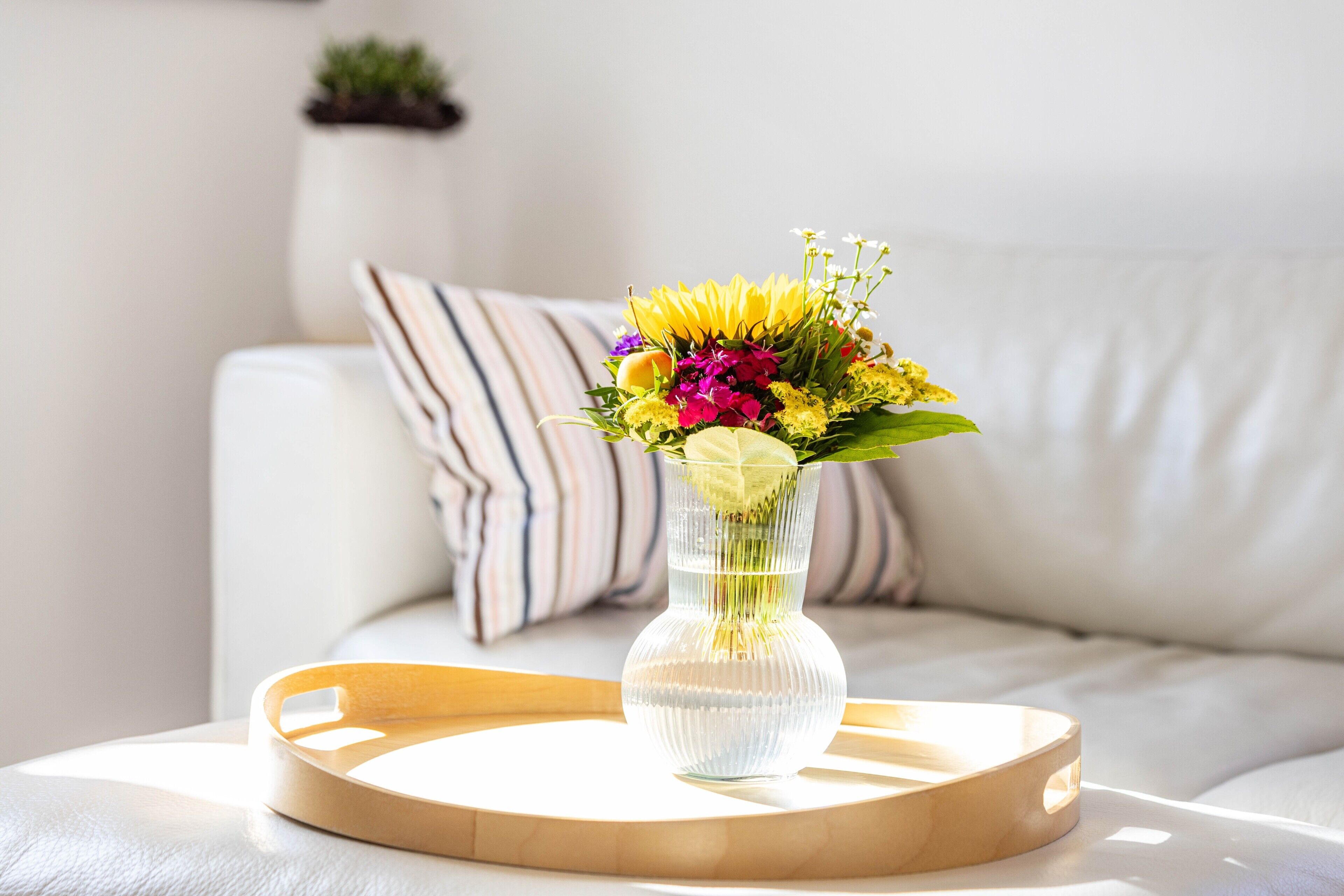 Ein freier Blick auf ein weißes Sofa mit gestreiften Kissen, eine klare Vase mit Blumen auf einem Holztablett und eine kleine Topfpflanze im Hintergrund.