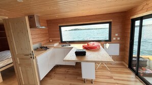 Een compacte keuken met een groot raam en uitzicht op het water, houten wanden en een klein eethoekje.