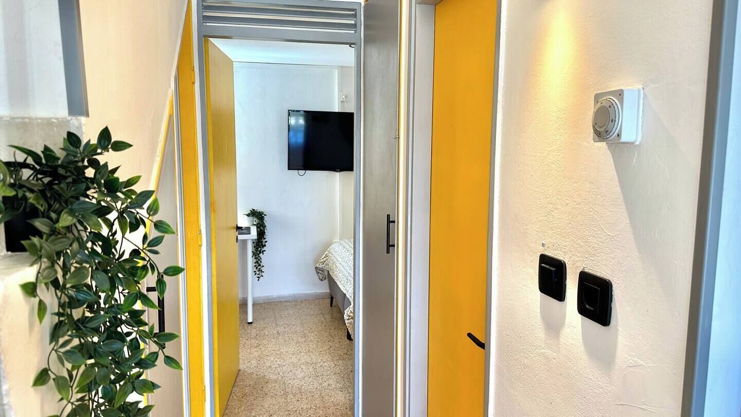 A hallway with a yellow door, a potted plant, a bed, a TV, and a wall-mounted light fixture.