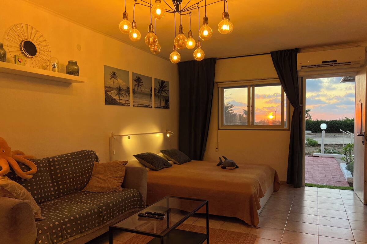 A cozy room with a sofa, a bed, a coffee table, and a nightstand. There are wall art, a ceiling light fixture, and a window with curtains.