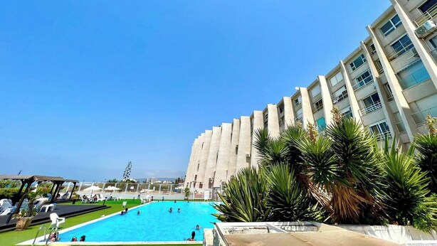 Una piscina rodeada por un edificio con varios balcones, un cielo azul despejado y algo de vegetación.