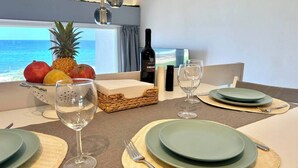 Una mesa de comedor dispuesta con platos, vasos y una botella de vino, con un frutero y vistas al océano.