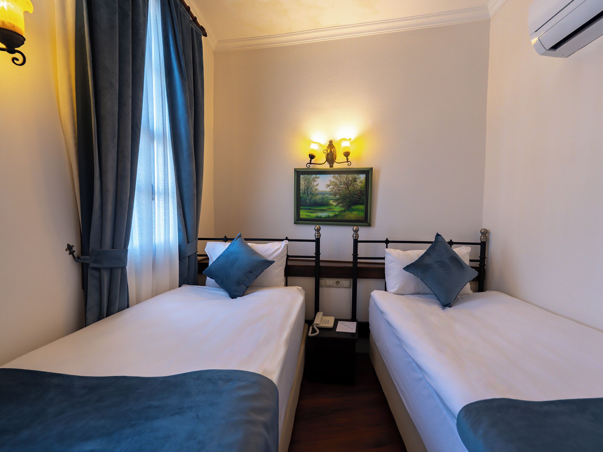 A small hotel room with two single beds, a nightstand with a telephone, and a painting on the wall.