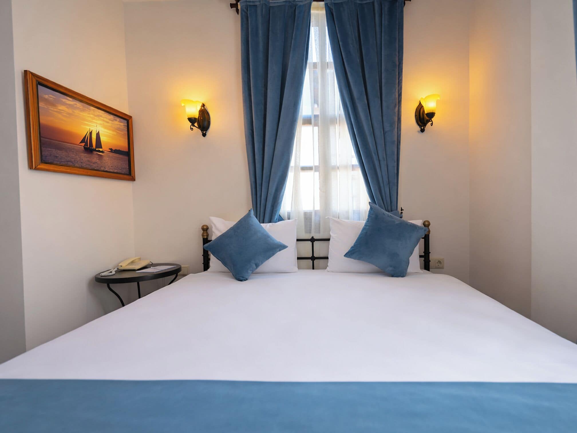 A hotel room with a large bed, two blue pillows, a bedside table with a lamp, a framed picture on the wall, and curtains.