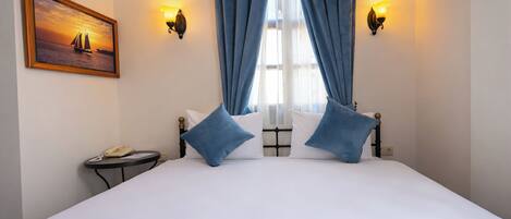 Habitación de hotel con una cama grande, dos almohadas azules, una mesita de noche con una lámpara, un cuadro enmarcado en la pared y cortinas.