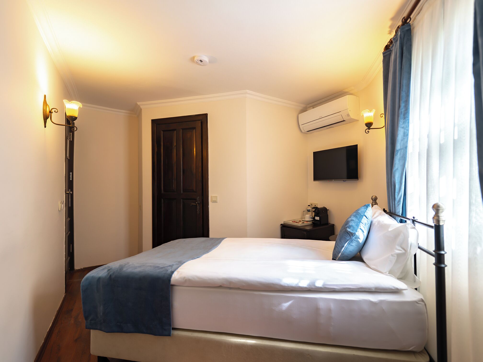 A hotel room with a bed, a flat-screen TV, an air conditioning unit, a bedside table with a lamp, and a door.