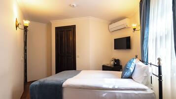 A hotel room with a bed, a flat-screen TV, an air conditioning unit, a bedside table with a lamp, and a door.