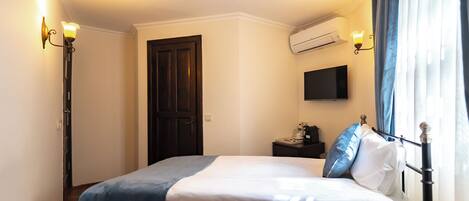 A hotel room with a bed, a flat-screen TV, an air conditioning unit, a bedside table with a lamp, and a door.