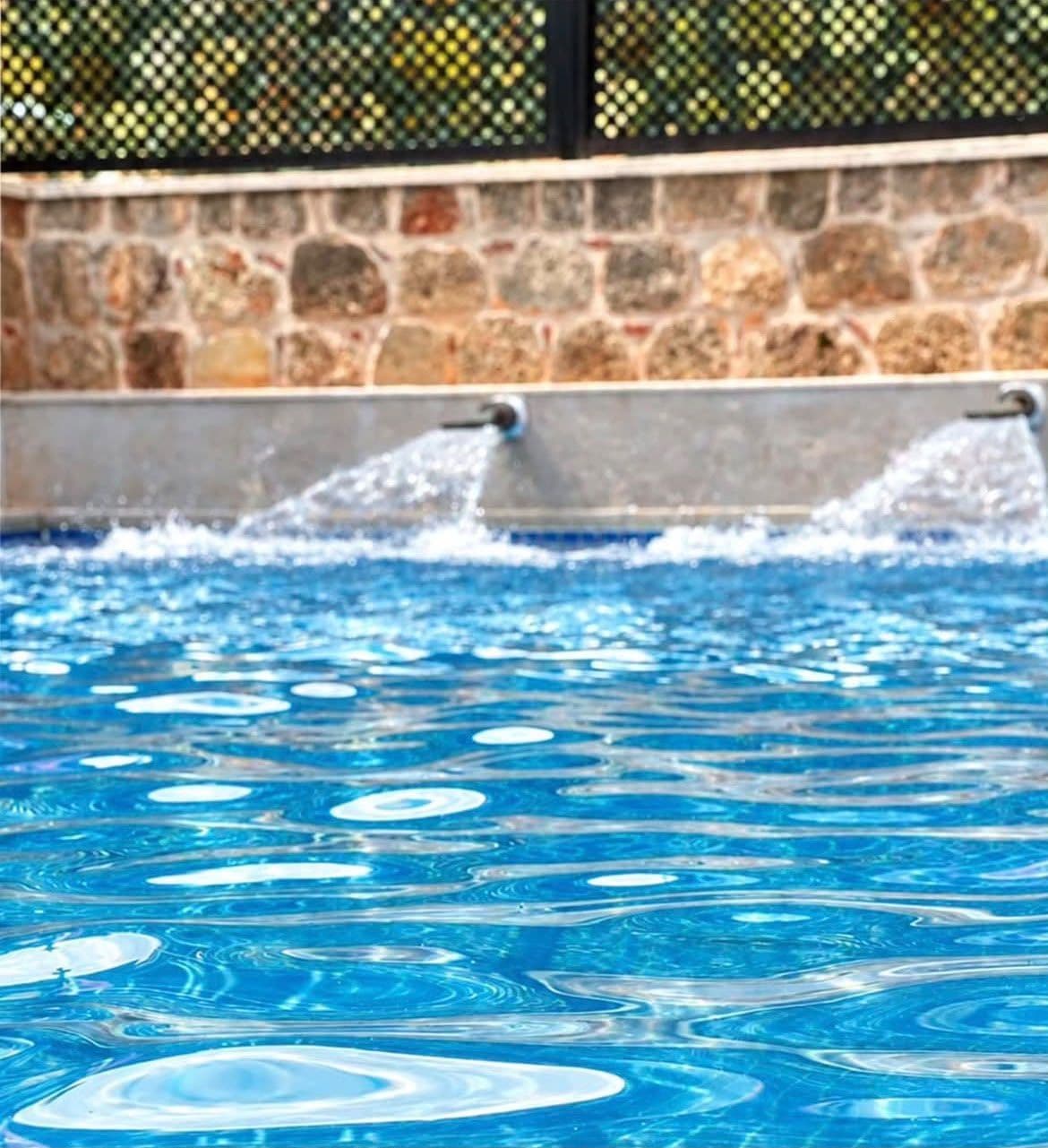 A swimming pool with water jets, surrounded by a stone wall and a metal fence.