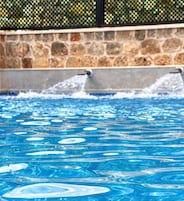 Una piscina con chorros de agua, rodeada por un muro de piedra y una valla de metal.