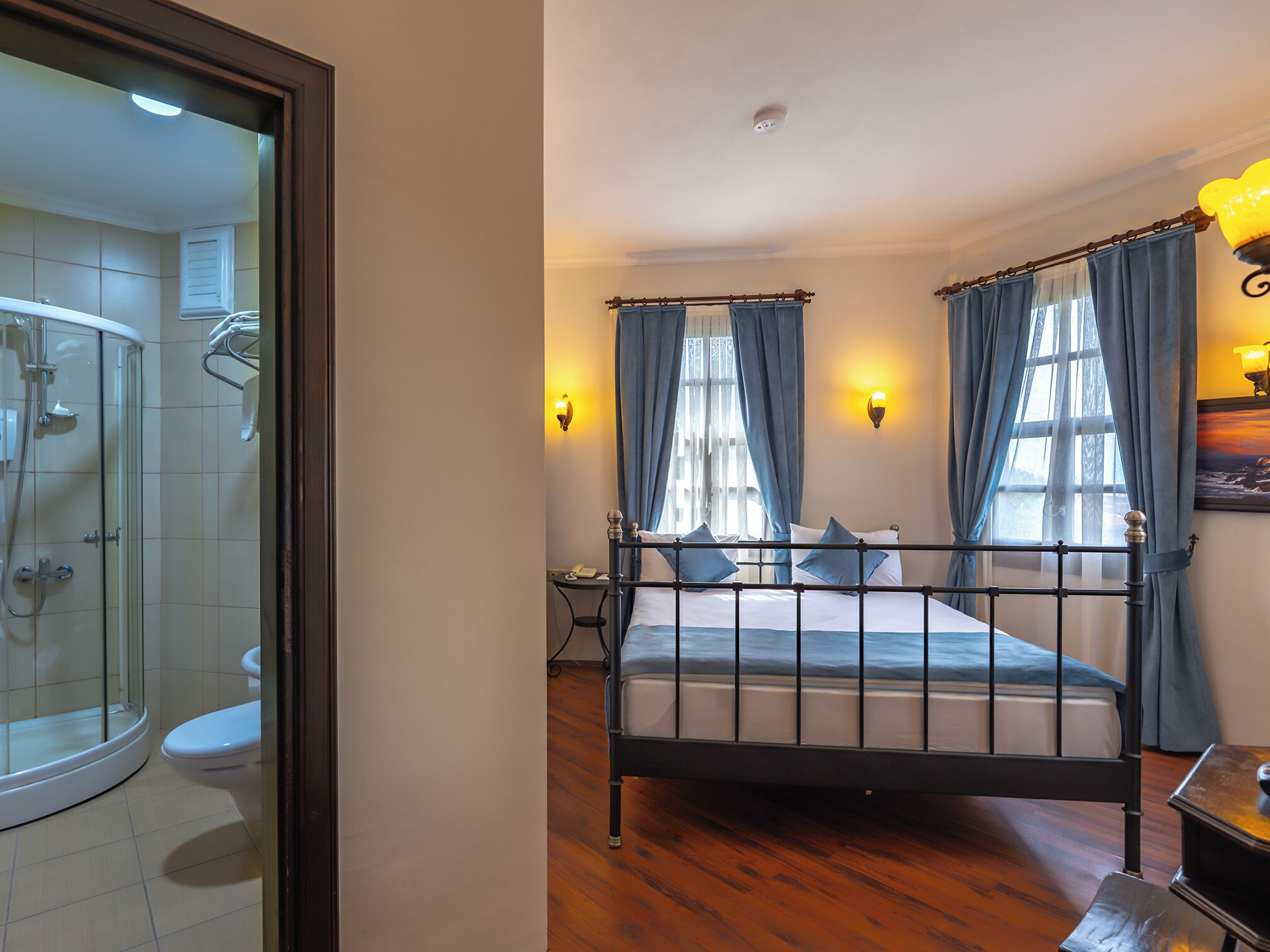 A bedroom with a bed, two bedside tables, a nightstand, a window with curtains, a wall-mounted light, and a bathroom with a shower and toilet.