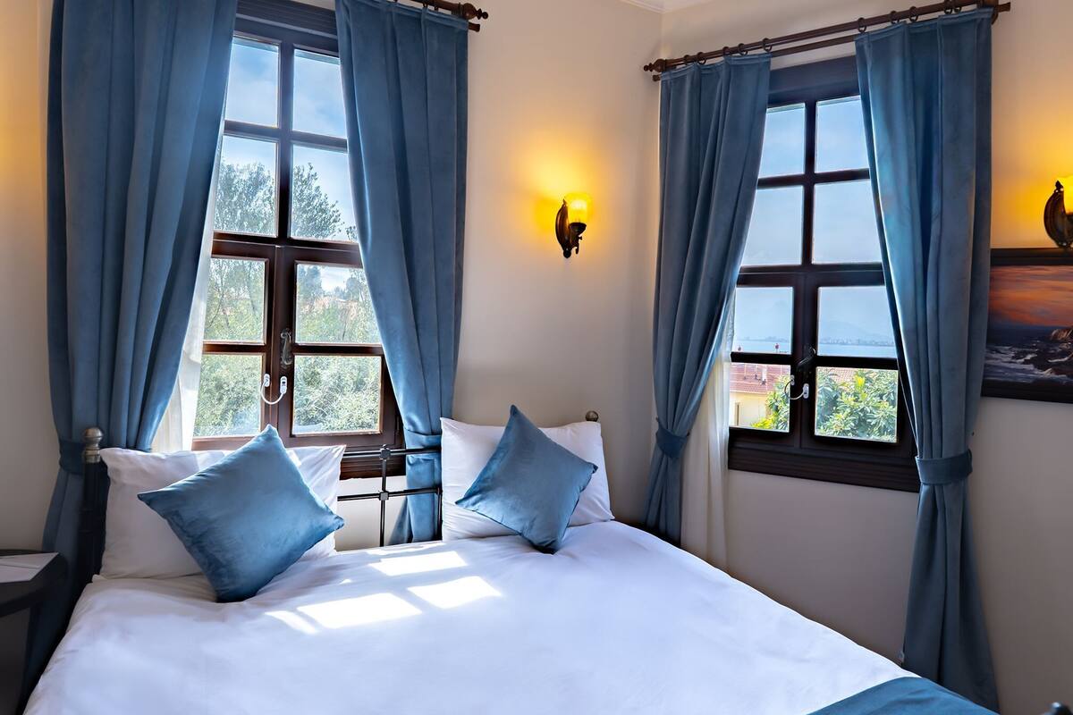 Ein Bett mit weißer Bettwäsche und blauen Kissen, ein Nachttisch, eine Wandlampe und ein Fenster mit Vorhängen.