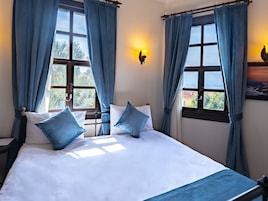 Una cama con sábanas blancas y almohadas azules, una mesita de noche, una lámpara de pared y una ventana con cortinas.