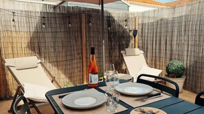 Un área de comedor al aire libre con dos sillones, una mesa preparada con platos, cubiertos y una botella de vino.