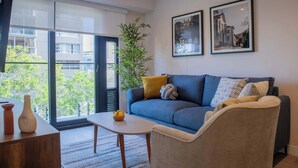 Un salón moderno con un sofá, un sillón y una pequeña mesa con jarrones. Hay un ventanal con vistas a edificios y árboles.