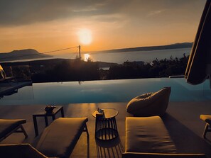 Ein Infinity-Pool mit Blick auf den Sonnenuntergang über dem Wasser, Liegestühle und ein kleiner Tisch.