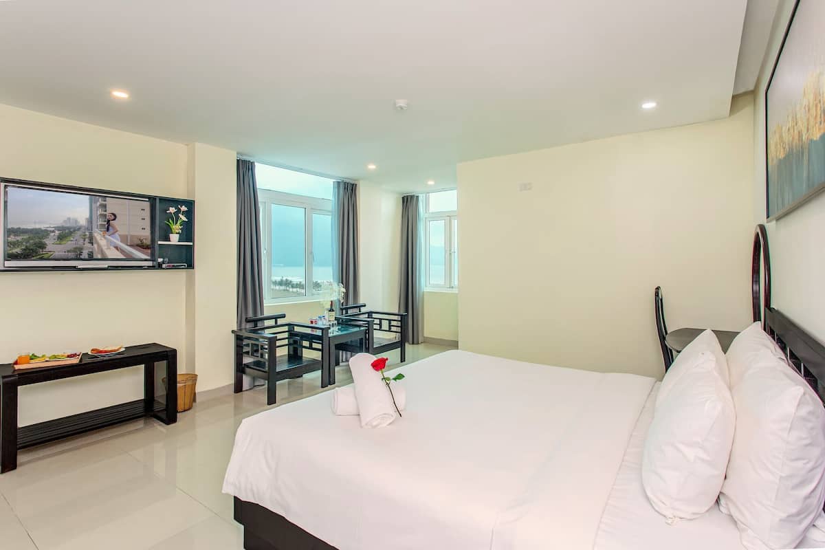 Habitación de hotel con cama, televisor montado en la pared, mesa de comedor con sillas y ventanas amplias con vistas.