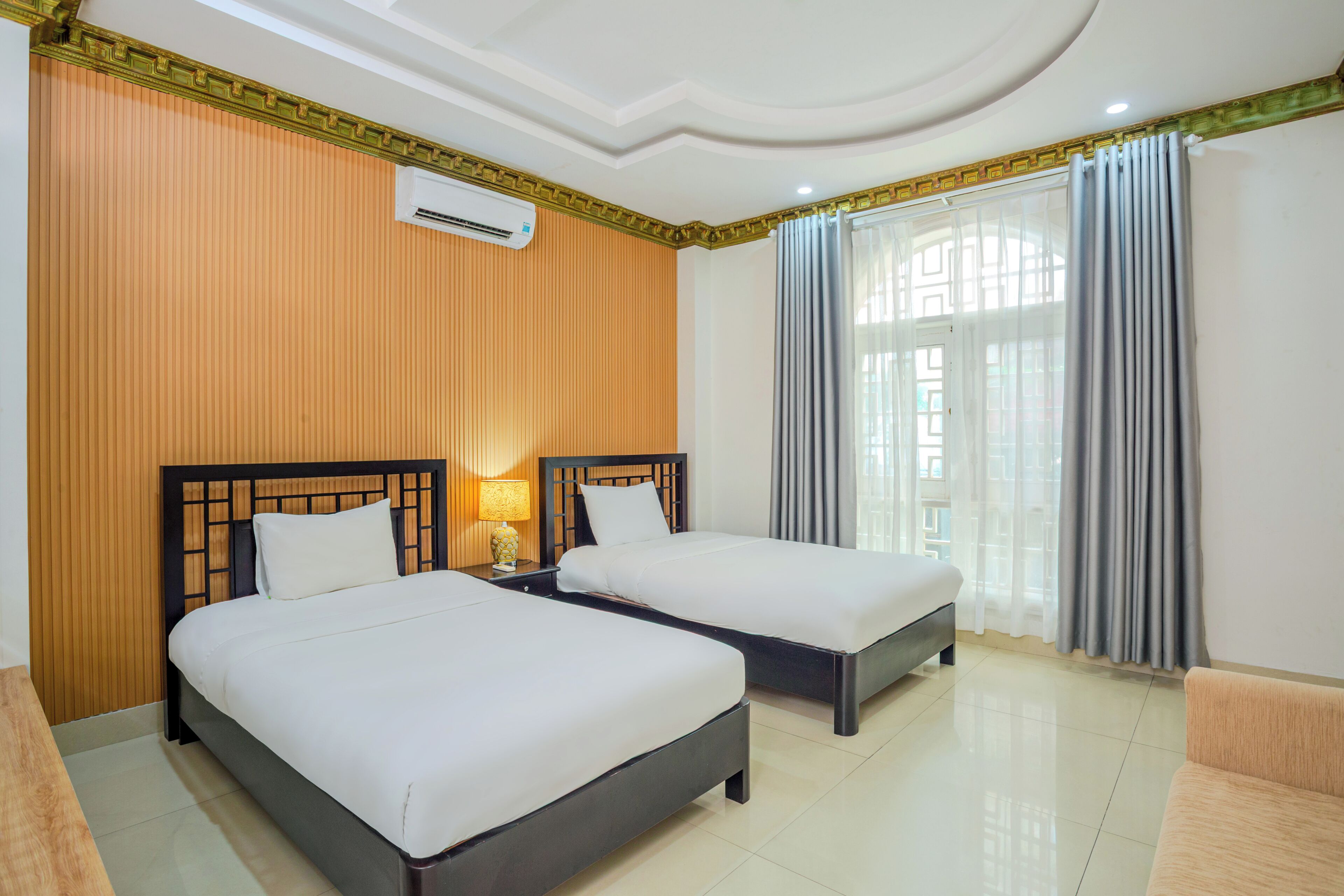 A hotel room with two beds, a wooden headboard, a nightstand with a lamp, and a large window with curtains.