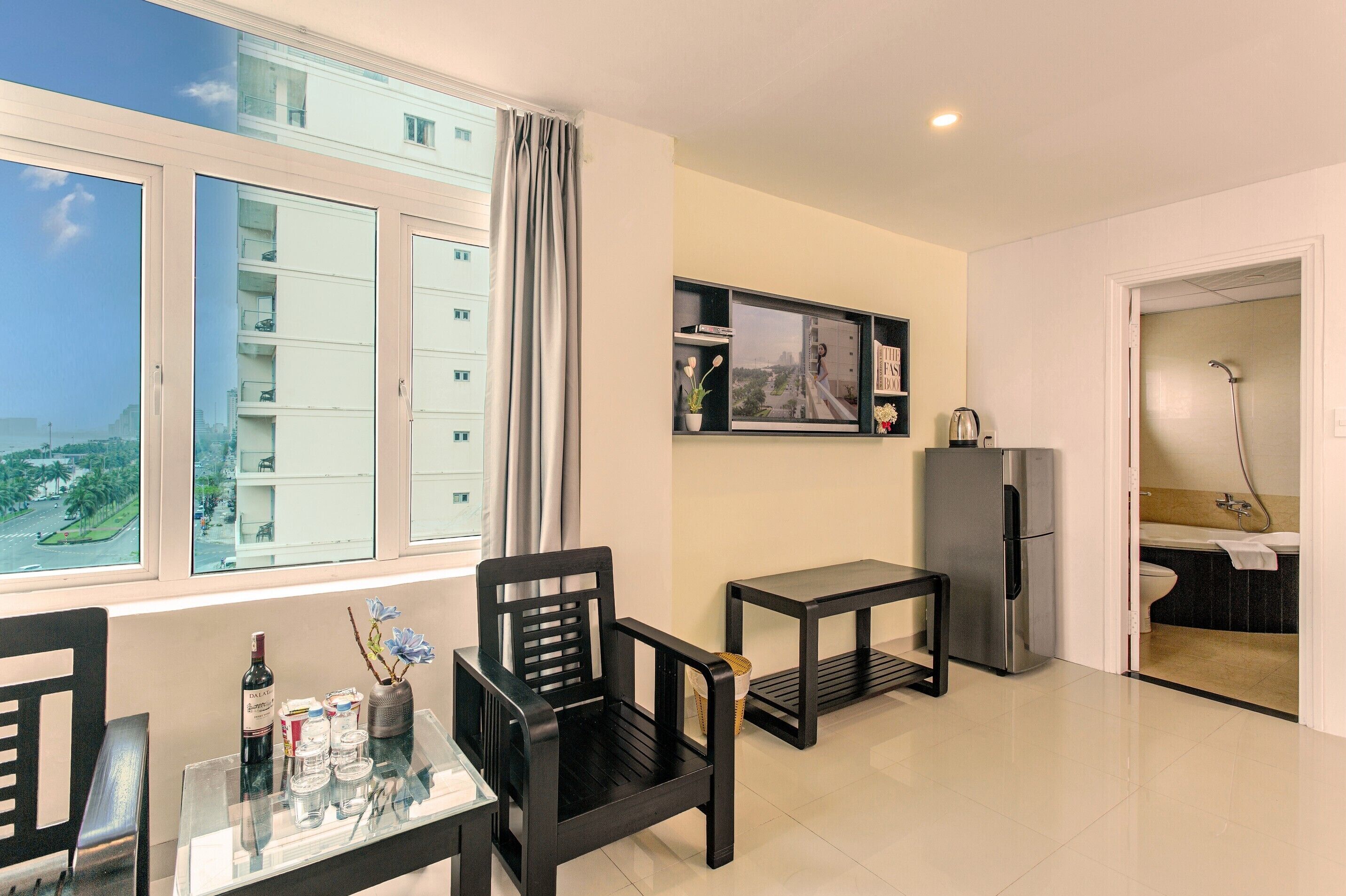 A modern living room with a large window, a dining table, two black chairs, a small black table, a refrigerator, and a view of a cityscape.