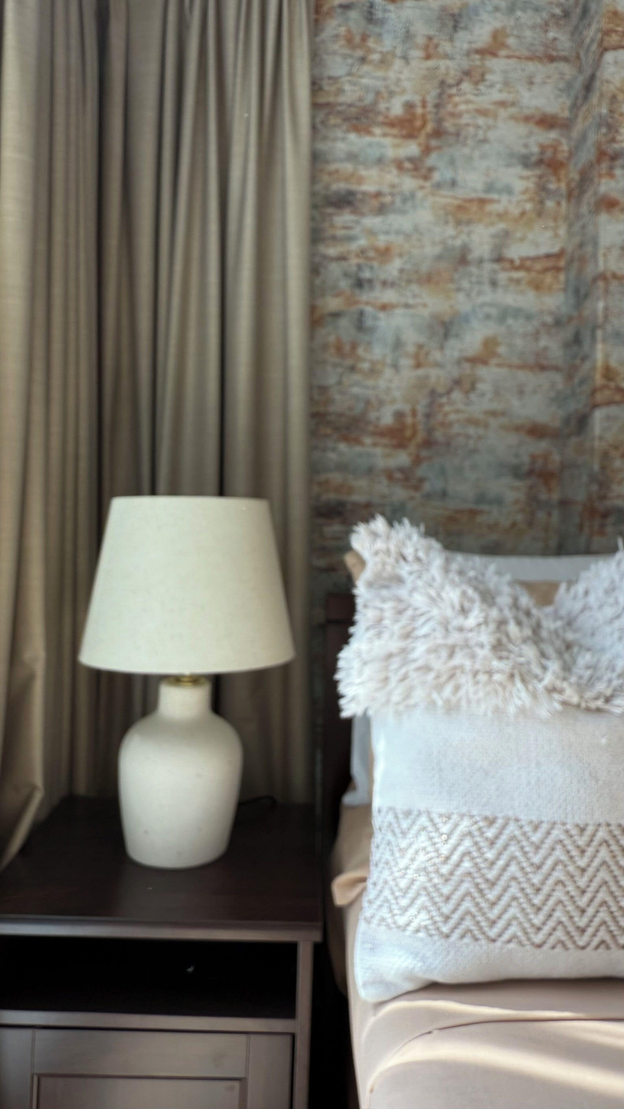 Un lit avec un oreiller blanc et une couverture à motifs, une lampe blanche sur une table de chevet, et un rideau en arrière-plan.