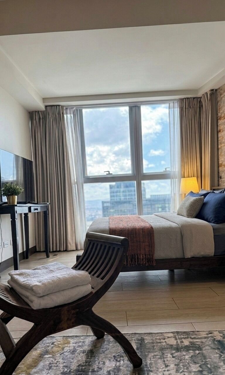 A hotel room with a bed, a desk with a TV, a chair, a large window with curtains, and a view of the city.