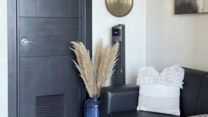 A modern living room with a dark wooden door, a blue vase with dried plants, a black sofa with a patterned throw, and a round wall decoration. - Luxury Uptown Parksuites - BGC (Taguig)
